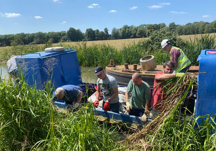 A team of Intertek colleagues on a small barge work on clearing vegetation on a river bank. 