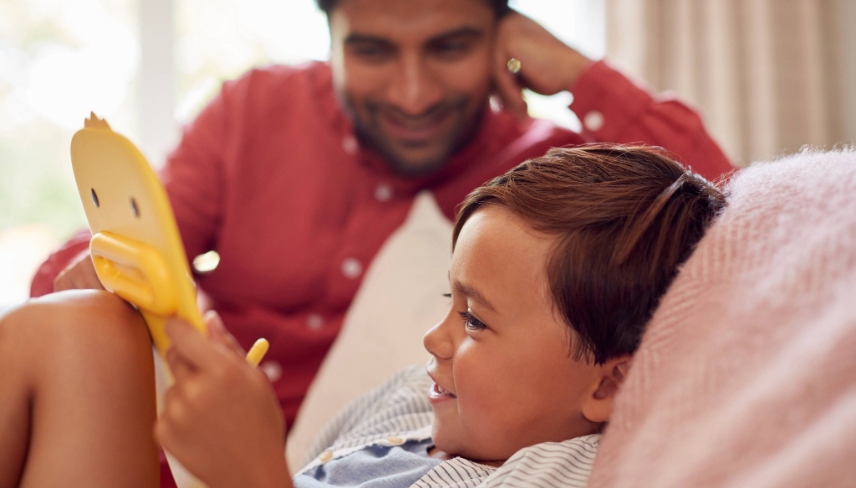 Father And Son At Home On Sofa In Lounge Playing With Digital Tablet Or Electronic Toy Together
