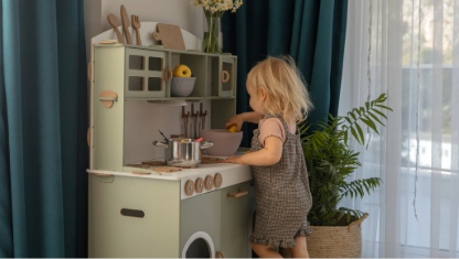 A young child smiles while playing with toy kitchen toys in a well-decorated, light-filled room.