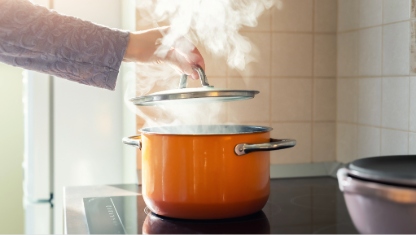Female hand open lid of enamel steel cooking pan on electric hob with boiling water or soup and scenic vapor steam backlit by warm sunlight at kitchen. 