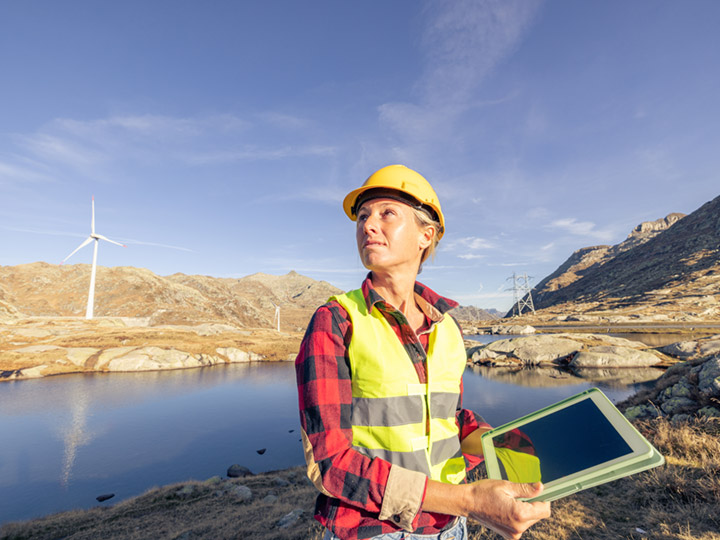 woman in protective gear standing, holding a tablet, in front of a lake that sits across from a wind turbine