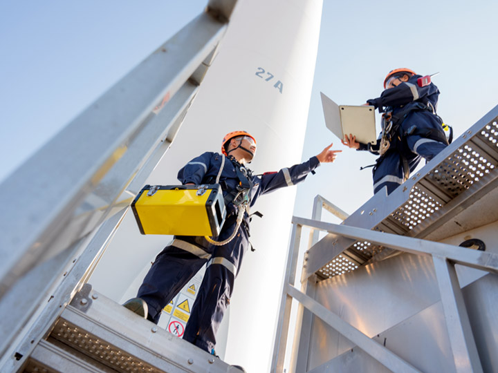 two workers stand atop a wind turbine