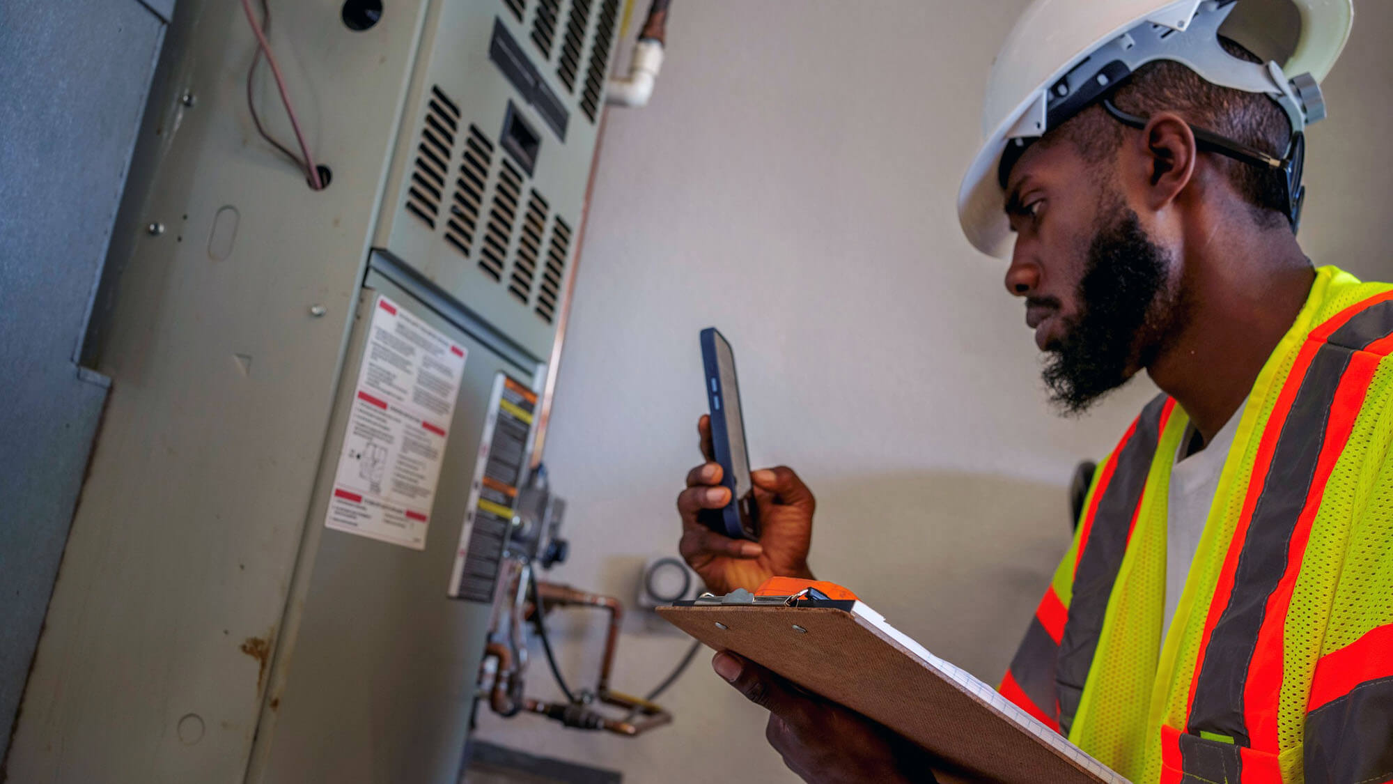 African American Man in His Twenties Photographing an Air Conditioning Unit in the Garage of a Residential Home Wearing Protective Workwear and a Hard Hat