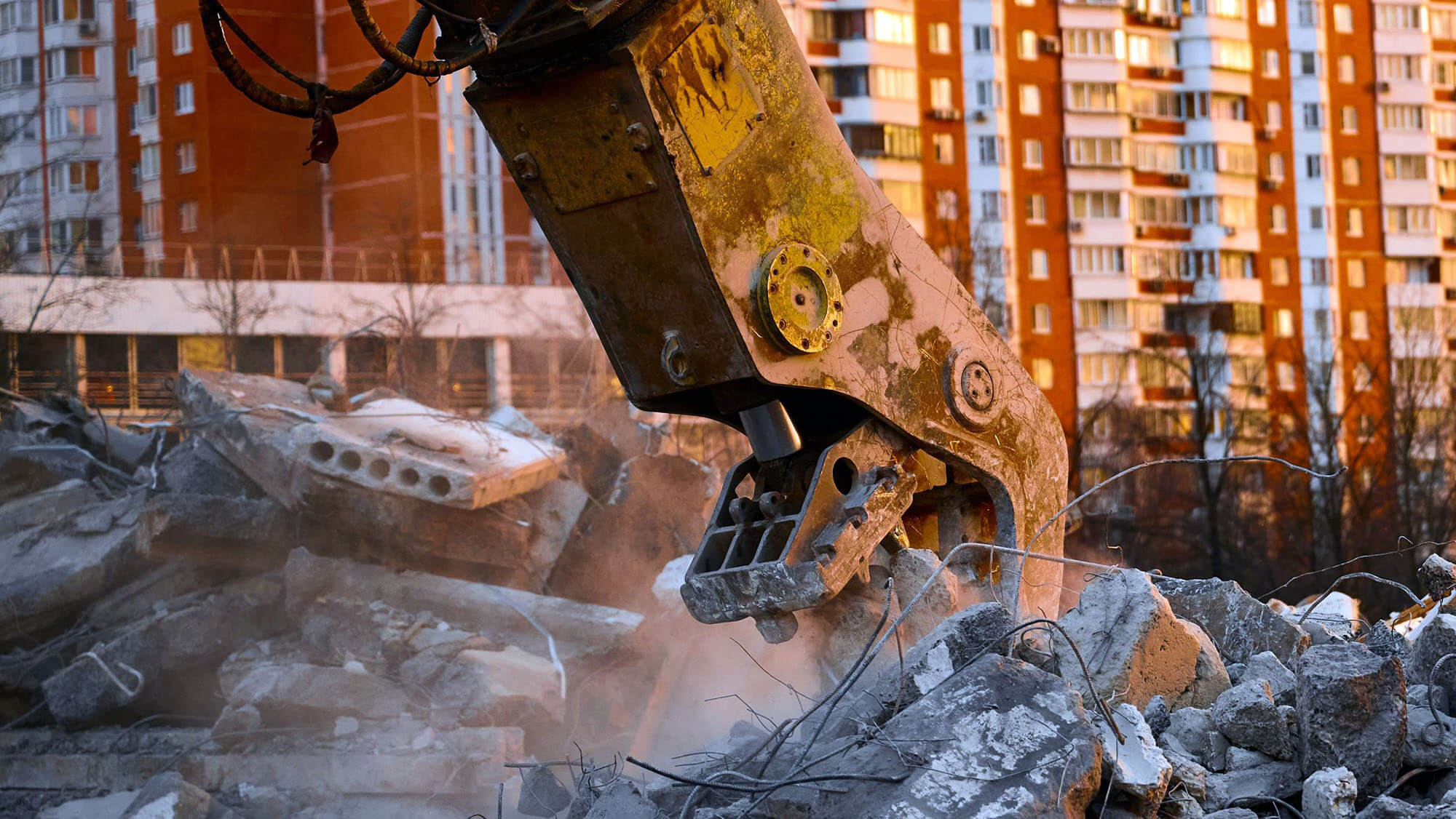 Concrete crusher on excavator rigging works with pile of reinforced cement leftovers for recycling at demolition site at sunset