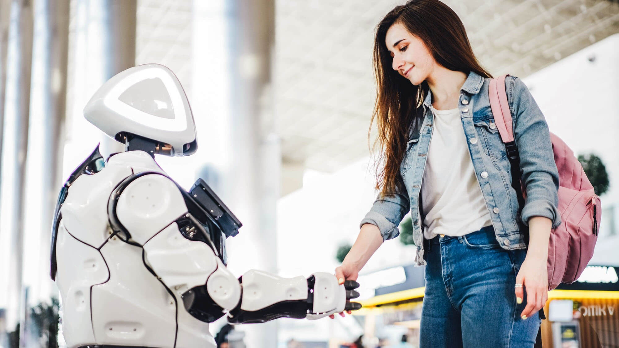 A young female traveler with a backpack smiles as she shakes hands with a sophisticated white humanoid robot in a bright, modern terminal. This interaction symbolizes the seamless integration of artificial intelligence and robotics into daily human life and travel experiences.