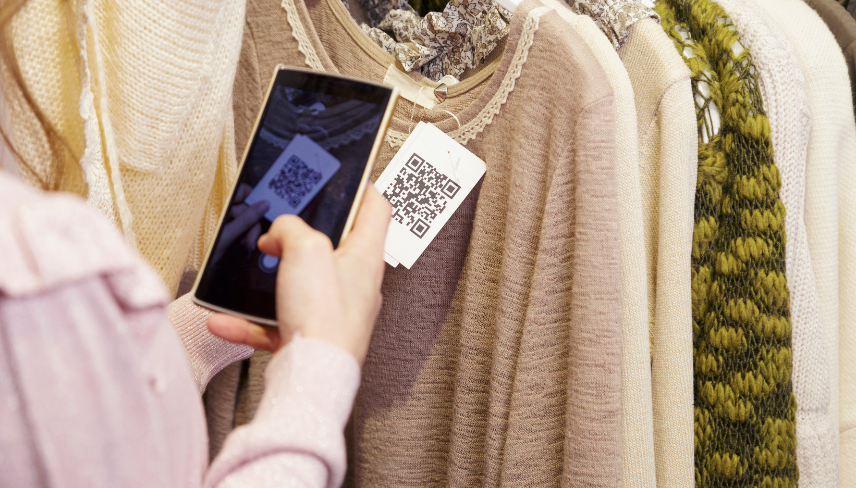 A person uses a smartphone to scan a QR code on a clothing tag while browsing sweaters on a store rack.